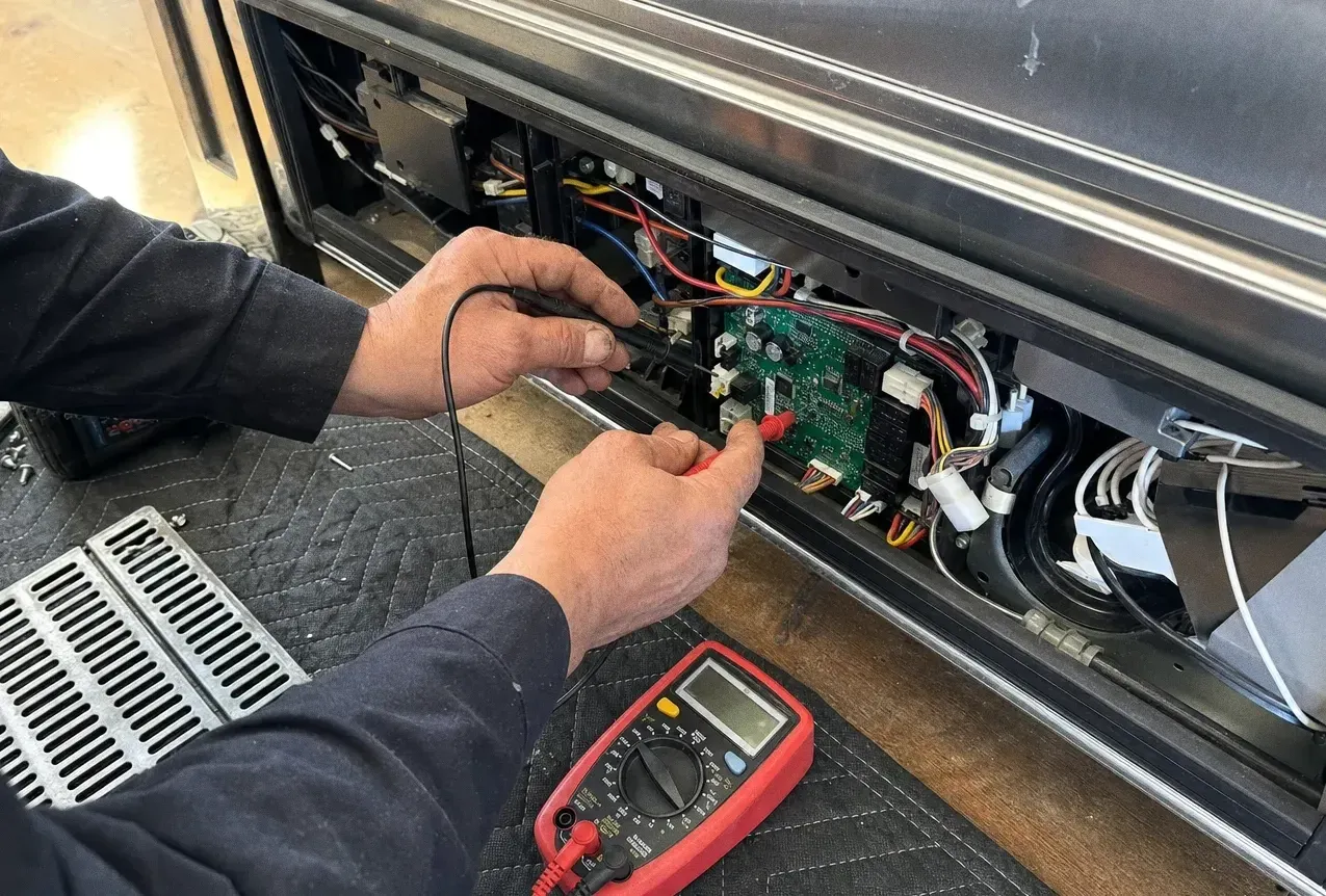 A multimeter probe held against a green circuit board inside the lower vent bay of a stainless built-in, meter resting on the grille