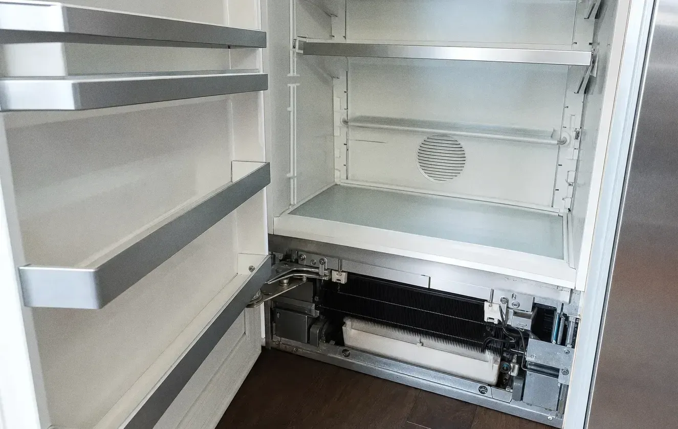 Empty white cabinet interior with glass shelves and the base service panel removed to show the vent behind it